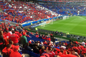 Eine große Menge Menschen in einem Stadion schaut ein Fußballspiel zu, mit Menschen auf Sitzen und Stehplätzen, Fahnen, einem Torpfosten und Deckenbeleuchtung unter einer elektrisierenden Atmosphäre.
