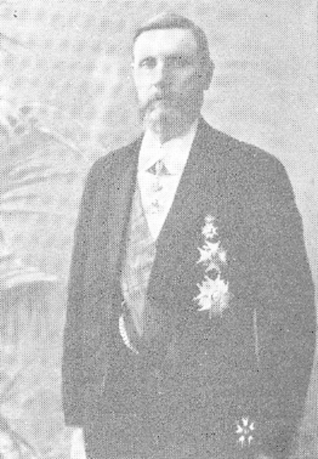 Ein Mann in einem schwarzen und weißen Anzug mit einem weißen Hemd und schwarzer Krawatte steht vor einer üppigen grünen Pflanze und hat einen ernsten Gesichtsausdruck.