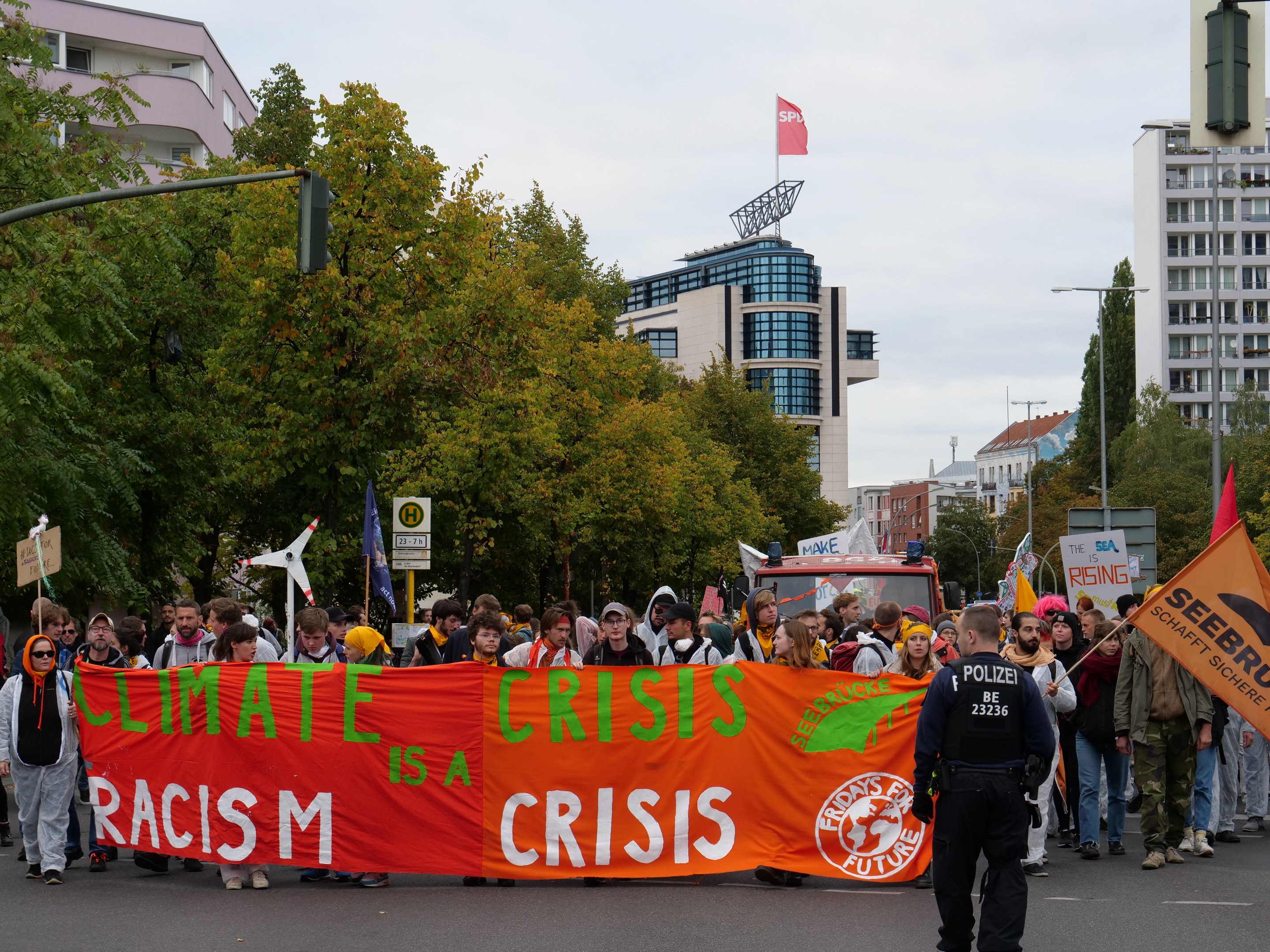 Gruppe von Menschen marschiert eine von Bäumen gesäumte Straße entlang und hält ein "Klima-Krise ist eine Krise"-Schild hoch, mit parkenden Fahrzeugen, Gebäuden und einem klaren Himmel im Hintergrund.