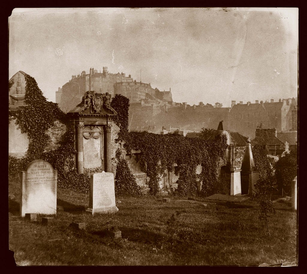 Altes Schwarz-Weiß-Foto eines Friedhofs mit Grabsteinen, Efeu und Gebäuden im Vordergrund und einer Burg im Hintergrund unter einem sichtbaren Himmel.