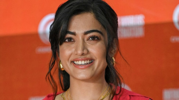 Eine Frau in einem roten Kleid mit einem eleganten Dutt, schwarzen High Heels und dezentem Make-up lächelt in die Kamera vor einem hellgelben Banner mit der Aufschrift 'Die besten Schönheits Looks von Sonam Kapoor beim Cannes Film Festival.'