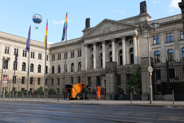 Großes modernes Gebäude mit vielen Fenstern, als Bundestag in Berlin identifiziert, umgeben von Straßeninfrastruktur, Fußgängern, Fahrzeugen und Bäumen unter einem bewölkten Himmel.