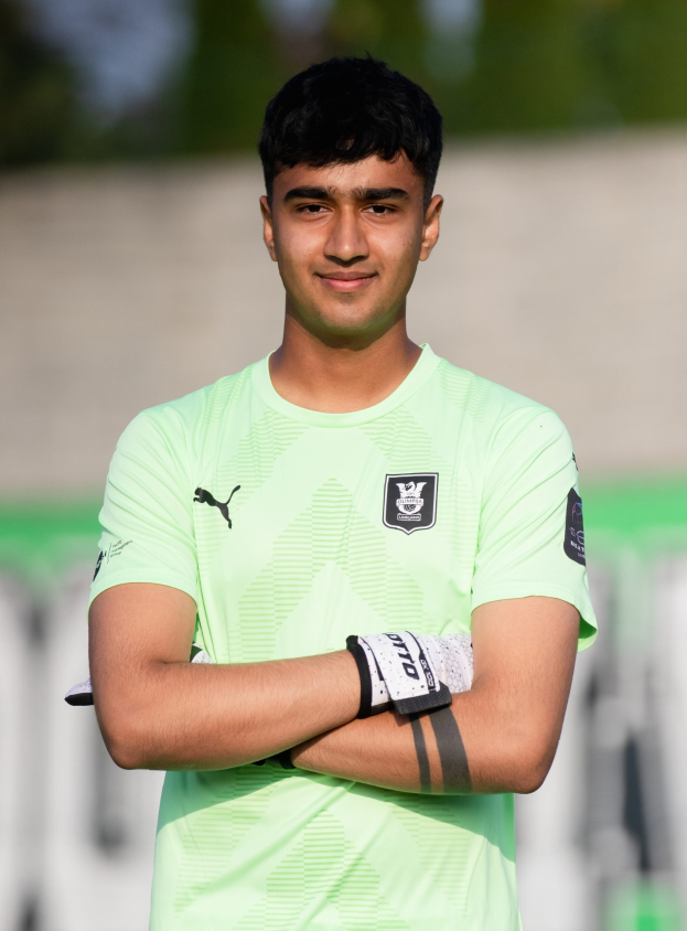 Ein Mann in einer blauen Jacke und Mütze steht auf einem Fußballfeld während eines Trainings, mit Bäumen und einem Tor im Hintergrund.