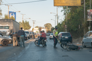 Eine Gruppe von Menschen steht um ein verunglücktes Motorrad am Straßenrand mit mehreren Fahrzeugen, darunter ein Lastwagen, und einer Hintergrundlandschaft aus Bäumen, Pfählen, Laternen und Schildern unter dem Himmel.