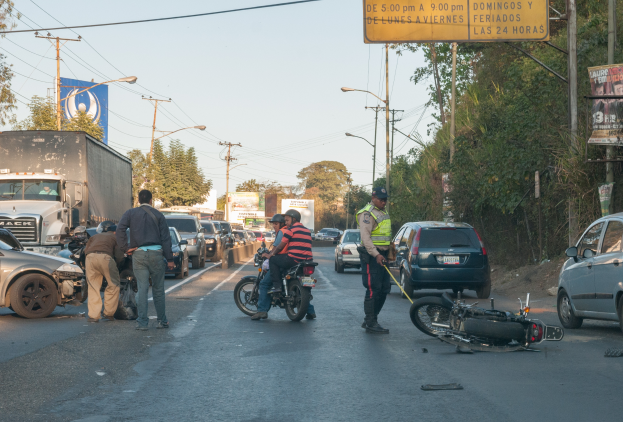 Eine Gruppe von Menschen steht um ein verunglücktes Motorrad am Straßenrand mit mehreren Fahrzeugen, darunter ein Lastwagen, und einer Hintergrundlandschaft aus Bäumen, Pfählen, Laternen und Schildern unter dem Himmel.