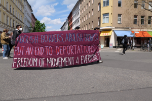 Eine Gruppe von Menschen marschiert auf der Straße und hält ein Transparent mit der Aufschrift "Abolish Borders, Abolish Frontiers, Put an End to Deportations, Freedom of Movement 4 Everyone" hoch, mit Gebäuden, Bäumen und Fahrrädern, die die Straße säumen, unter einem bewölktem Himmel.