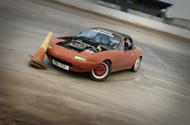 Eine Person fährt ein rotes Auto auf einer Rennstrecke mit einem Verkehrskegel vorneweg, der Hintergrund ist unscharf und zeigt hohe Geschwindigkeit.