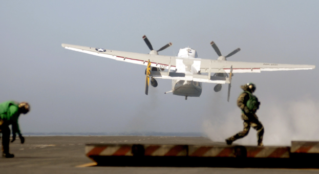 Ein Flugzeug startet von einem Flugzeugträger, mit zwei Personen, die auf der Rollbahn darunter gehen und dem Himmel im Hintergrund.