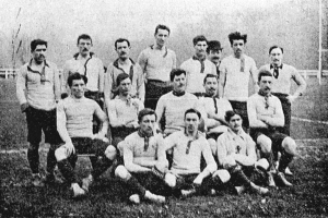 Ein Schwarz-Weiß-Foto einer Fußballmannschaft aus den 1920er Jahren, mit einigen stehenden und anderen auf dem Boden sitzenden Spielern, sowie Pfählen, Bäumen und einem klaren Himmel im Hintergrund.