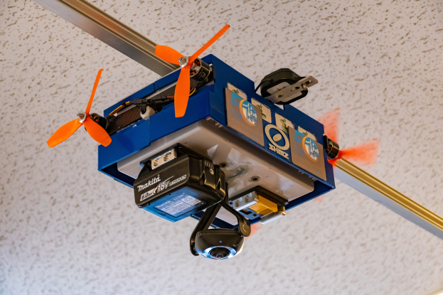 Ein kleiner blauer Drohne mit orangen Propellern fliegt in der Luft, befestigt an einer Metallstange an der Decke, wahrscheinlich für die Hausautomatisierung verwendet.