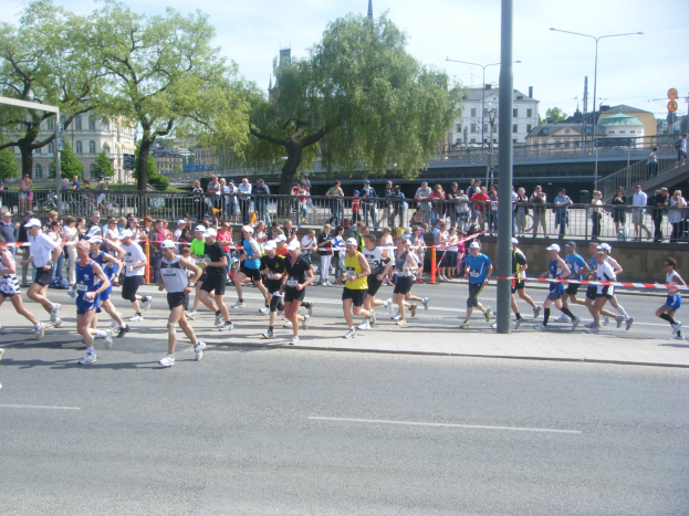 Gruppe von Läufern beim Marathon auf einer Straße mit Zielpfosten und Band, umgeben von Metallzaun und Absperrungen, Zuschauern auf dem Gehweg, Pfosten, Schilder, Brücke, Gebäuden, Bäumen und bewölktem Himmel.