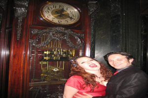 Ein Paar, das zusammen steht und lächelt, mit einer Uhr und einer Wand im Hintergrund.