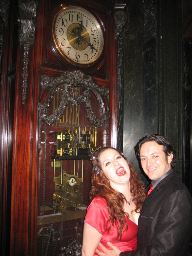 Ein Paar, das zusammen steht und lächelt, mit einer Uhr und einer Wand im Hintergrund.