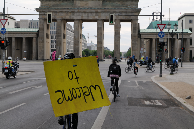 Eine Gruppe von Radfahrern in Helmen fährt auf einer Straße am Brandenburger Tor in Berlin, Deutschland vorbei, wobei einer ein gelbes Schild hält, Lichtmasten, Verkehrszeichen, Gebäude, Bäume und einen klaren blauen Himmel im Hintergrund.