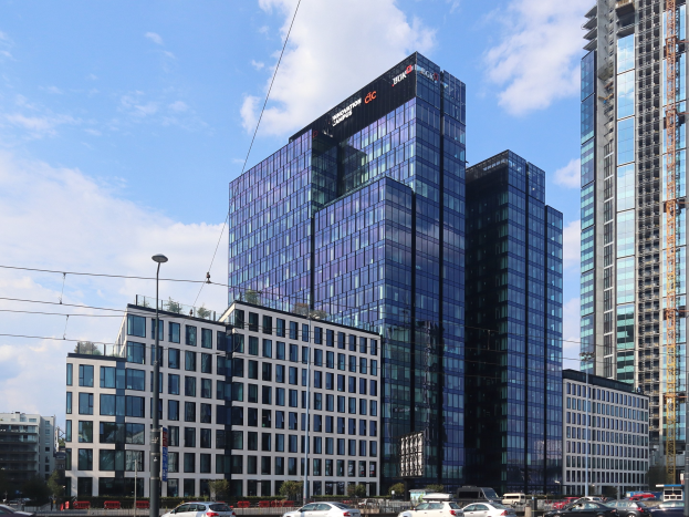 Außenansicht des Deutschen Bank-Hauptquartiers in Frankfurt, Deutschland, mit umgebender städtischer Infrastruktur einschließlich Straßenlaternen, Fahrzeugen und Bäumen.