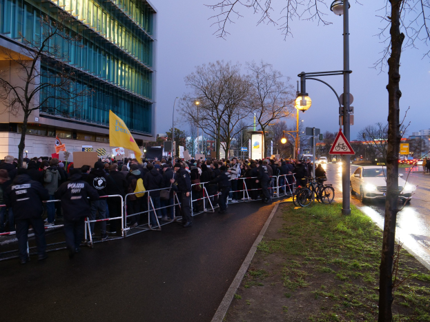 Eine große Gruppe von Menschen mit Schildern steht vor einem Gebäude mit Barrikaden und Bäumen, was auf eine Demonstration in Berlin hinweist.