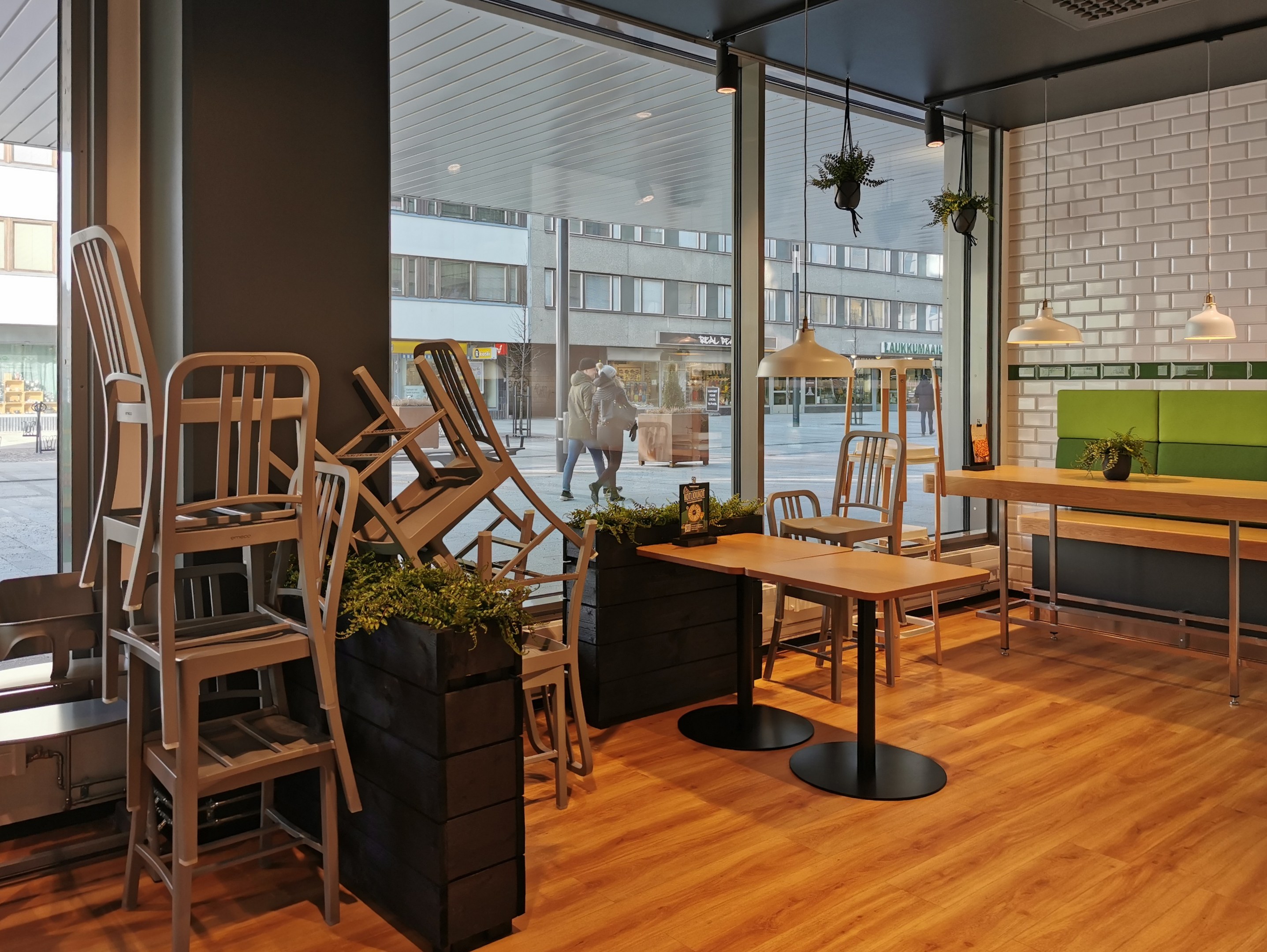 Ein Berliner Restaurant mit Holztischen und -stühlen in der Nähe eines großen Fensters, das eine Außenstraße mit Fußgängern, Gebäuden und städtischen Elementen zeigt.