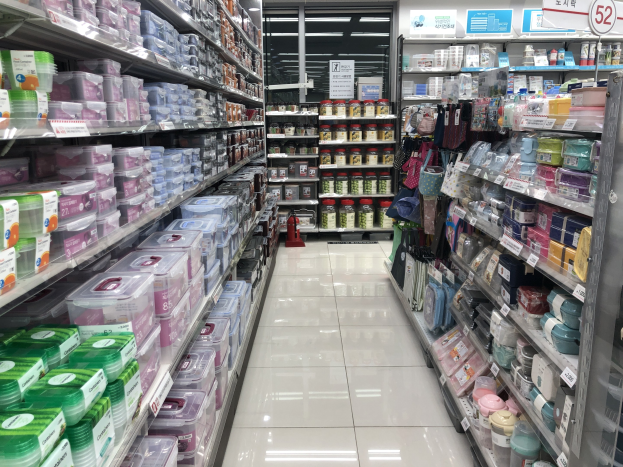 Ein unordentlicher chinesischer Supermarktgang mit Regalen voller Kartons, Flaschen, Tüten und anderen Artikeln, mit beleuchteten Schildern, Deckenlampen und einem sichtbaren Feuerlöscher auf dem Boden.