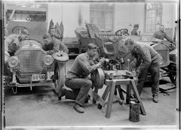 Eine Gruppe uniformierter Männer, die ein Auto in einer schwarz-weißen Garage reparieren, mit Werkzeugen, die auf einem zentralen Tisch verstreut sind.