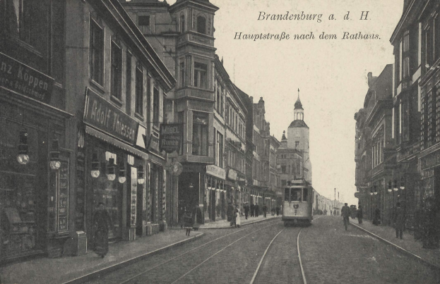 Ein altes Schwarz-Weiß-Foto einer Stadtstraße in Brandenburg, Deutschland, mit einer Tram auf Schienen, Menschen auf dem Gehweg, Gebäuden mit Fenstern, Schildern und Straßenlaternen unter einem bewölkten Himmel.