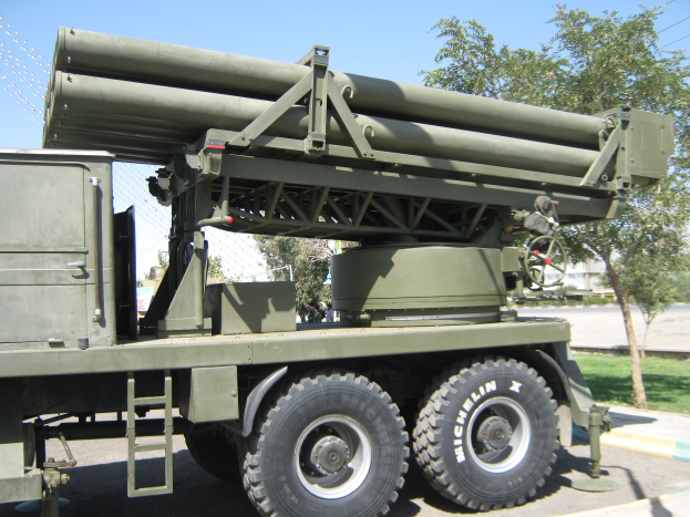 Militärlastwagen transportiert eine Rakete auf einer Straße, die von Gras, Bäumen, Gebäuden, einem Maschendrahtzaun und einem klaren blauen Himmel gesäumt ist.