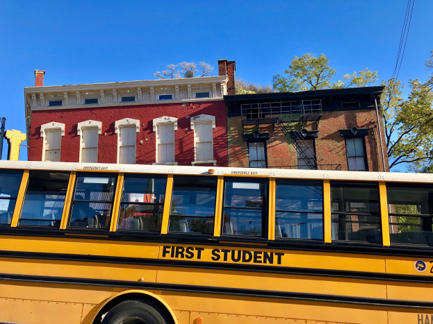 Gelber Schulbus mit der Aufschrift "First Student" vor einem roten Backsteingebäude mit einigen Menschen drinnen, Bäumen und einem klaren blauen Himmel im Hintergrund geparkt.
