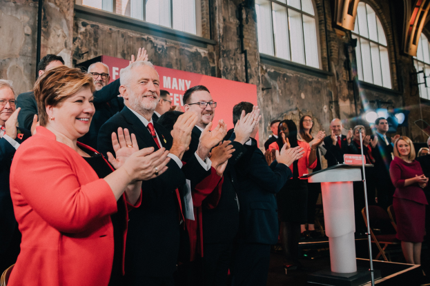 Eine Gruppe von Menschen, die vor einem Publikum klatschen, mit einem Podium, einem Mikrofon und einer Tafel mit Text auf der rechten Seite sowie Stühlen, einer Fahne, einer Wand, Fenstern und Lichtern im Hintergrund.