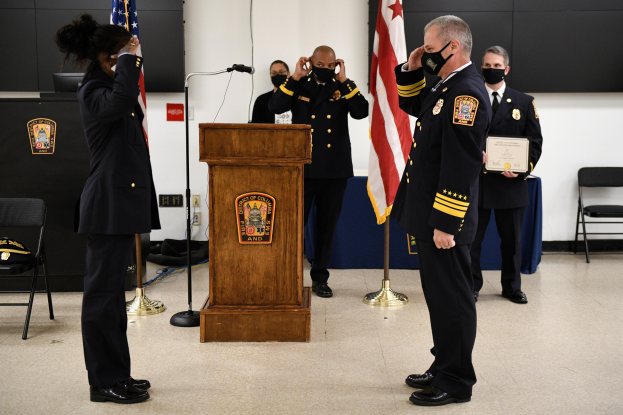 Gruppe uniformierter Personen bei einer Vereidigung eines neuen Feuerchefs, der eine Urkunde hält, mit Flaggen, Podium und maskierten Teilnehmern im Hintergrund.