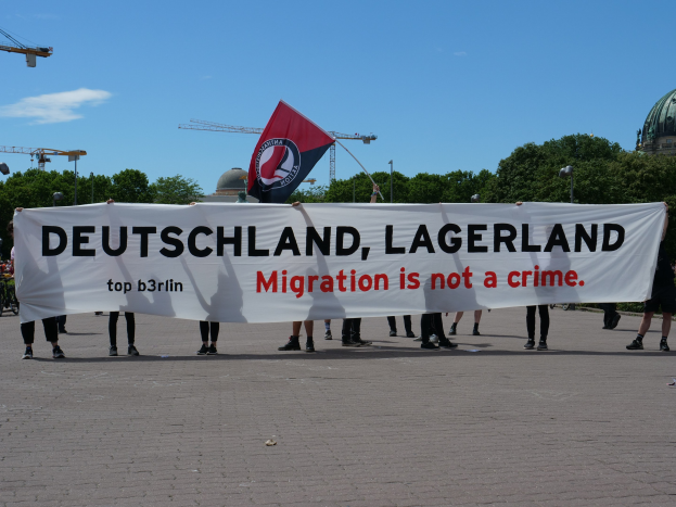 Menschen mit einem Transparent mit der Aufschrift "Deutschland, Lagerland Migration ist kein Verbrechen" und einer Flagge, mit Straßenlaternen, Bäumen, Kränen, einem Gebäude und einem bewölkten Himmel im Hintergrund.