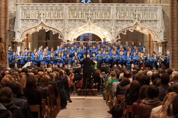 Eine große Gruppe von Menschen steht vor einem Chor in einer Kirche. Die Chormitglieder sitzen auf Bänken und einige stehen, und eine Person in der Mitte hält ein Mikrofon mit Ständer; dahinter ist eine Wand mit Skulpturen und einem Glasfenster, und links im Bild sind Statuen zu sehen.