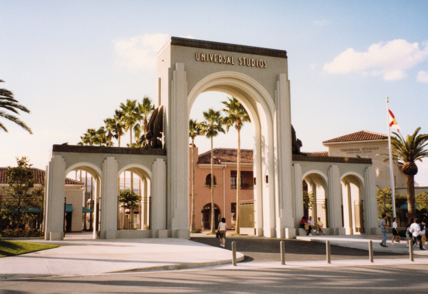 Eingang zu Universal Studios Hollywood mit einem Bogen mit Text, einer Statue, Besuchern, Grünflächen, Gebäuden, einem Fahnenmast mit einer Flagge und einem bewölkten Himmel.