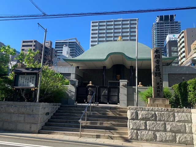 Großes Gebäude mit grüner Dachfläche in städtischer Umgebung mit umliegenden Treppen, Geländern und Grünflächen, betrachtet über eine Straße mit Hochhäusern und einem klaren Himmel im Hintergrund.