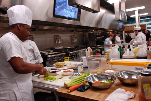 Eine Gruppe von Köchen in einer Küche bei der Zubereitung von Essen auf einem Schneidbrett, mit einem Koch im Vordergrund in einer weißen Schürze und Mütze, der ein Messer hält und von verschiedenen Utensilien, Schüsseln, Gemüse, einem Handy, Flaschen und anderen Gegenständen umgeben ist, mit einem Fernseher und Deckenlampen im Hintergrund.