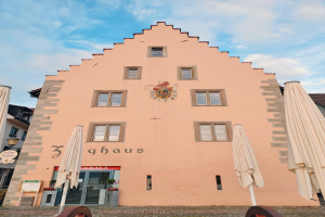 Ein pinkes Gebäude mit einem zentralen Uhrenturm, umgeben von Fenstern, Schirmen, einem Schild, einer Straßenlaterne, Topfpflanzen, unter einem bewölkten Himmel, repräsentiert München, Deutschland.