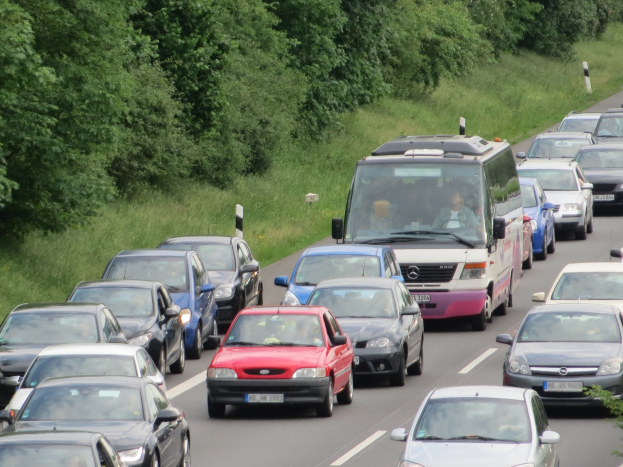 Ein Stau auf einer Autobahn mit vielen Autos und einem Van, Menschen in den Fahrzeugen sichtbar, Bäume und Gras im Hintergrund.