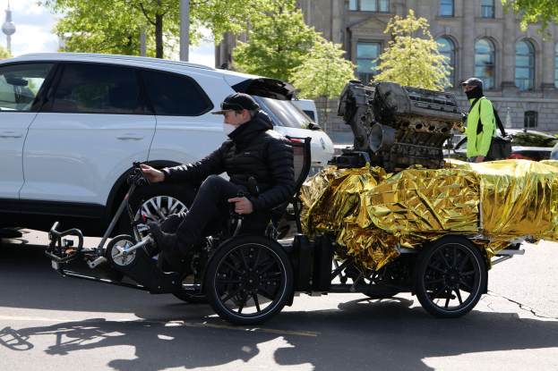Ein Mann im Rollstuhl mit einem großen Motor am Rücken, der ein Objekt hält, auf einer Straße umgeben von Fahrzeugen, Bäumen, Gebäuden, Polen und einem klaren blauen Himmel.