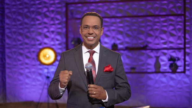 Stephen Colbert, Moderator von *The Daily Show with Trevor Noah*, steht auf einer Bühne und hält ein Mikrofon in der Hand, trägt einen Anzug, Krawatte und Brille, mit einem Scheinwerfer und Ständer im Hintergrund.