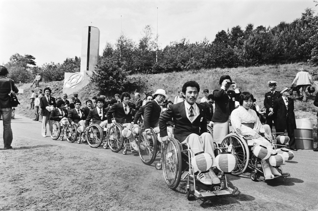 Eine Gruppe von Menschen im Rollstuhl steht an der Straße, trägt verschiedene farbige Kleider und einige Mützen, mit Bäumen, einem Turm und einem klaren blauen Himmel im Hintergrund, dargestellt in Schwarz-Weiß.