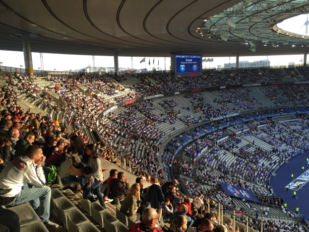 Große Zuschauermenge in einem Stadion bei einem Fussballspiel, mit einer Bühne auf der rechten Seite, Fahnen, Masten, einem Bildschirm und der Allianz Arena in München, Deutschland im Hintergrund.