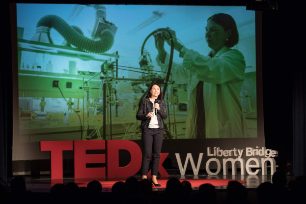 Frau auf einer Bühne mit Mikrofon, hinter ihr ein Display mit einem Bild einer Frau und Text, die eine Menge anspricht.