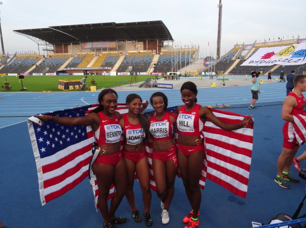 Vier Frauen in Sportkleidung stehen auf einer Laufbahn, lächeln und halten amerikanische Flaggen, mit einem Stadion und Himmel im Hintergrund.