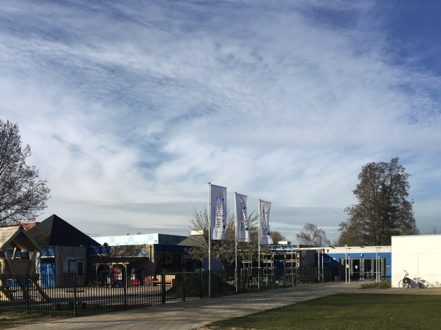 Großes Gebäude mit Metallzaun, Rasenfläche, Weg, Bäumen, Bannern, einem abgestellten Fahrrad, nahen Häusern und einem bewölkten Himmel, identifiziert als neuer Spielplatz der Schule.