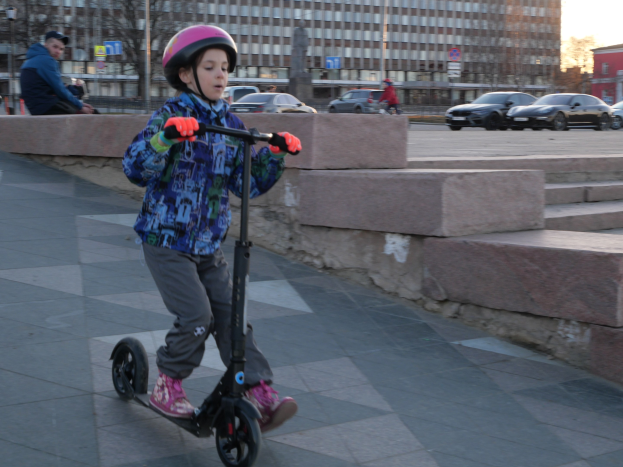 Ein junger Junge in Helm und Handschuhen fährt auf einem Roller eine Treppe auf dem Gehweg hinunter, mit Autos, Menschen, Bäumen, Pfosten, Brettern, Gebäuden und einem klaren blauen Himmel im Hintergrund.