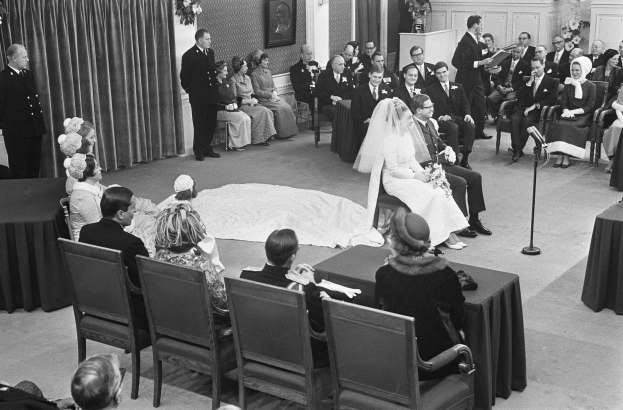 Schwarzes und weißes Foto einer Hochzeitszeremonie mit sitzenden und stehenden Menschen, ein Mann mit Mikrofon, eine Frau mit einem Strauß, ein Podium mit Mikrofon, Blumenarrangements, ein Vorhang, ein gerahmtes Foto an der Wand und eine Tür im Hintergrund.