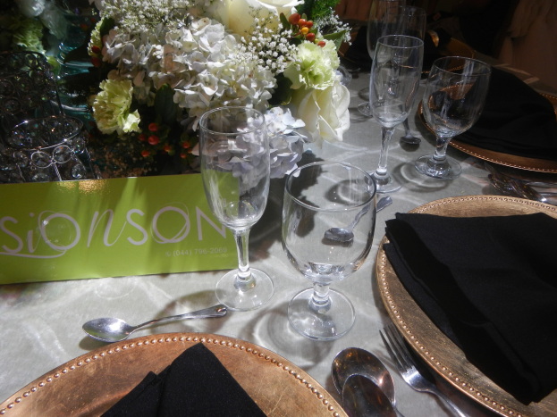 Ein formell gedeckter Esstisch mit goldenen Untersetzern, schwarzen Servietten, Besteck, Gläsern und einem Blumenbouquet, begleitet von einer Texttafel.
