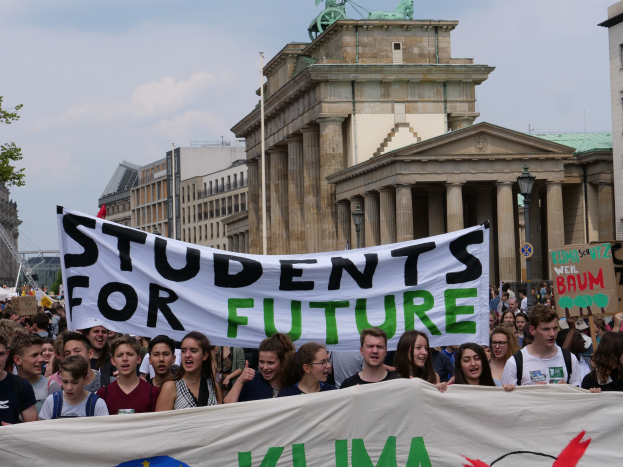 Eine Gruppe von Schülern marschiert in Berlin, eine buntfarbene "Students for Future"-Fahne schwingend vor einem Hintergrund aus Gebäuden, Bäumen und Himmel.