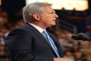 Ein Mann im Anzug und mit Krawatte steht an einem Podium mit zwei Mikrofonen, sieht ernst aus und hat einen unscharfen Hintergrund.