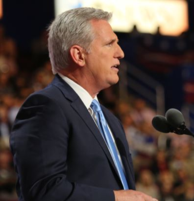 Ein Mann im Anzug und mit Krawatte steht an einem Podium mit zwei Mikrofonen, sieht ernst aus und hat einen unscharfen Hintergrund.