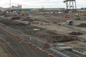 Eine Baustelle mit einer Bahnstrecke, umgeben von einem Metallzaun, mit mehreren Fahrzeugen, Verkehrskegeln, Rohren und anderen Gegenständen, mit Laternenmasten, Gebäuden und einem bewölktem Himmel im Hintergrund.