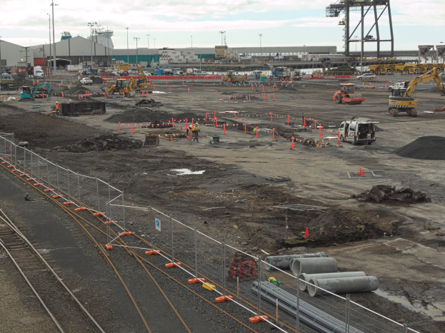 Eine Baustelle mit einer Bahnstrecke, umgeben von einem Metallzaun, mit mehreren Fahrzeugen, Verkehrskegeln, Rohren und anderen Gegenständen, mit Laternenmasten, Gebäuden und einem bewölktem Himmel im Hintergrund.